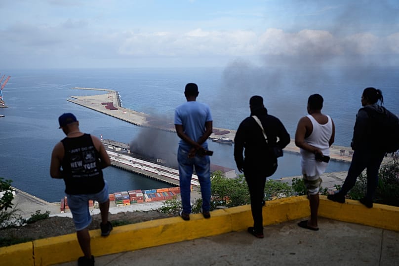 Men watch smoke rising from a dock after explosions were heard at La Guaira port, Venezuela, Saturday, Jan. 3, 2026